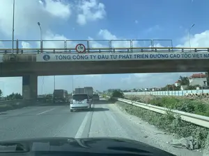 Bối cảnh là một con đường cao tốc tại Việt Nam, được chụp từ góc nhìn bên trong một chiếc ô tô đang di chuyển. Phía trước là một cây cầu vượt, trên đó có dòng chữ lớn màu xanh "TỔNG CÔNG TY ĐẦU TƯ PHÁT TRIỂN ĐƯỜNG CAO TỐC VIỆT NAM" và logo của công ty. Trên lan can cầu có một biển báo hình tròn viền đỏ ghi giới hạn chiều cao là "4,75m". Nhiều loại ô tô đang di chuyển trên đường. Bầu trời quang đãng với mây trắng. Lề đường bên phải có đất, cỏ mọc và một số ống cống bê tông, cho thấy khu vực này có thể đang được xây dựng. Xa hơn là một vài tòa nhà dân cư.。（日本語訳）。舞台はベトナムのある高速道路で、移動中の車内からの視点で撮影されています。前方には陸橋があり、その上には「TỔNG CÔNG TY ĐẦU TƯ PHÁT TRIỂN ĐƯỜNG CAO TỐC VIỆT NAM」という大きな青い文字と、その会社のロゴがあります。橋の欄干には、高さ制限「4.75m」と書かれた赤枠の円形標識があります。道路上では様々な種類の車が走行しています。空は晴れ渡り、白い雲が浮かんでいます。右側の路肩には土や草が生え、コンクリート製の土管がいくつかあり、この地域が建設中である可能性を示しています。さらに遠くには、いくつかの住宅ビルが見えます。