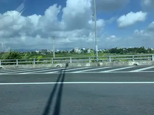 Bức ảnh được chụp từ một phương tiện đang di chuyển, nhìn ra ngoài qua cửa sổ. Cảnh vật cho thấy một con đường cao tốc hoặc một cây cầu có lan can bê tông và rào chắn kim loại. Trên mặt đường nhựa có các vạch kẻ đường màu trắng và bóng của một vật thể dài (có thể là cột đèn) đổ xuống. Phía bên kia lan can là một khung cảnh nông thôn với những cánh đồng xanh, cây cối (như cây chuối) và các ngôi nhà dân. Ở phía xa, có thể thấy một số tòa nhà đang được xây dựng và một dãy núi mờ ảo ở đường chân trời. Bầu trời trong xanh với những đám mây trắng lớn, cho thấy thời tiết đẹp.。（日本語訳）。写真は移動中の乗り物から窓の外を撮影したものです。アスファルトの路面には白い車線があり、長い物体（おそらく街灯）の影が落ちています。欄干の向こう側は、緑の田畑、木々（バナナの木など）、そして民家のある田園風景です。遠くには、建設中の建物がいくつか見え、地平線にはぼんやりとした山脈があります。空は青く澄み、大きな白い雲が浮かんでおり、天気が良いことを示しています。