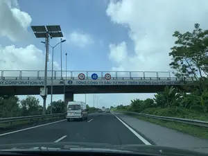 Bức ảnh được chụp từ bên trong một chiếc ô tô đang di chuyển trên một tuyến đường cao tốc ở Việt Nam. Con đường có nhiều làn xe và lan can bảo vệ ở hai bên. Phía trước có một số phương tiện khác đang lưu thông, nổi bật là một chiếc xe van màu trắng. Băng qua phía trên con đường là một cây cầu vượt, trên đó có dòng chữ lớn màu xanh: "TỔNG CÔNG TY ĐẦU TƯ PHÁT TRIỂN ĐƯỜNG CAO TỐC VIỆT NAM". Ngoài ra, trên cầu còn có các biển báo giao thông: biển báo tốc độ tối đa 120 km/h, biển báo tốc độ tối thiểu 60 km/h và biển báo giới hạn chiều cao 4,75m. Bên lề đường có các cột đèn chiếu sáng và một cột lớn được trang bị các tấm pin năng lượng mặt trời và camera giám sát. Cảnh quan hai bên đường là cây cối xanh tươi, bao gồm cả những bụi chuối. Bầu trời trong xanh và có nhiều mây trắng.。（日本語訳）。写真はベトナムの高速道路を走行中の車内から撮影されたものです。道路には複数の車線があり、両側にはガードレールが設置されています。前方には他の車両が数台走行しており、中でも白いバンが目立ちます。道路の上を横切るように陸橋があり、そこには「TỔNG CÔNG TY ĐẦU TƯ PHÁT TRIỂN ĐƯỜNG CAO TỐC VIỆT NAM」（ベトナム高速道路投資開発総公社）という大きな青い文字が書かれています。さらに、橋には交通標識もあります。最高速度120km/hの標識、最低速度60km/hの標識、そして高さ制限4.75mの標識です。路肩には街灯のポールと、ソーラーパネルや監視カメラが設置された大きなポールがあります。道路の両側の景観は緑豊かな木々で、バナナの茂みも含まれています。空は青く澄んでおり、白い雲が多く浮かんでいます。