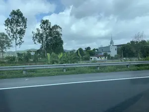 Bức ảnh ghi lại khung cảnh nông thôn nhìn từ một phương tiện đang di chuyển trên đường cao tốc. Ở tiền cảnh là mặt đường nhựa và dải hộ lan kim loại. Phía sau dải hộ lan là cảnh vật làng quê với nhiều cây cối, bao gồm các cây cao và những bụi cây chuối. Ở phía xa, có thể nhìn thấy một số ngôi nhà và một nhà thờ Công giáo nổi bật với tháp chuông cao theo kiến trúc Gothic. Xa hơn nữa là những ngọn đồi xanh mờ. Bầu trời nhiều mây. Không có văn bản hay chữ viết nào có thể nhận dạng được trong ảnh.。（日本語訳）。写真は、高速道路を移動中の乗り物から見た田園風景を記録したものです。前景には、アスファルトの路面と金属製のガードレールがあります。ガードレールの後ろには、高い木々やバナナの木の茂みなど、多くの木々がある村の風景が広がっています。遠くには、いくつかの家と、ゴシック建築の高い鐘楼が特徴的なカトリック教会が見えます。さらにその先には、緑に霞んだ丘々があります。空は曇っています。写真には、認識できるテキストや文字はありません。