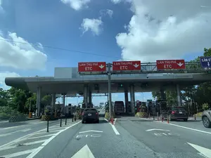 Hình ảnh chụp tại một trạm thu phí trên đường cao tốc vào một ngày trời trong xanh, có mây. Đây là trạm thu phí tự động không dừng (ETC). Phía trên các làn xe có nhiều biển báo lớn màu đỏ với dòng chữ 'LÀN THU PHÍ ETC' và mũi tên chỉ xuống. Trên mặt đường cũng có sơn chữ 'ETC' để chỉ dẫn các phương tiện. Nhiều loại xe ô tô, bao gồm xe con và xe khách, đang di chuyển qua các làn để trả phí. Có thể thấy các thiết bị của hệ thống thu phí như camera và barie tự động. Các đèn tín hiệu màu xanh lá cây có chữ 'XE CON', 'XE KHÁCH', 'XE TẢI' để phân loại phương tiện được phép đi vào làn.。（日本語訳）。青空が広がり、雲のある日に高速道路の料金所で撮影された画像。これはノンストップ自動料金収受システム（ETC）の料金所です。車線の上には、「LÀN THU PHÍ ETC」（ETC料金レーン）という文字と下向きの矢印が書かれた大きな赤い看板が多数あります。路面にも車両を誘導するために「ETC」の文字がペイントされています。乗用車やバスを含む多くの種類の自動車が、料金を支払うためにレーンを通過しています。カメラや自動遮断機といった料金収受システムの設備が見えます。緑色の信号機には、「XE CON」（乗用車）、「XE KHÁCH」（バス）、「XE TẢI」（トラック）という文字があり、レーンへの進入が許可されている車両を分類しています。