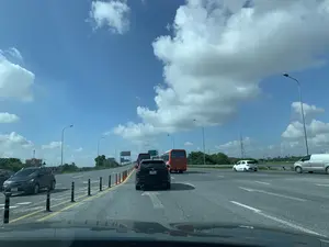 Bức ảnh được chụp từ bên trong một chiếc ô tô, nhìn ra một con đường cao tốc nhiều làn xe vào một ngày nắng. Bầu trời trong xanh với nhiều mây trắng. Trên đường có nhiều phương tiện đang tham gia giao thông, bao gồm một chiếc ô tô SUV màu đen ở giữa, một chiếc xe khách màu cam, một chiếc ô tô con màu xám ở bên trái, và một chiếc ô tô con màu trắng cùng một xe tải van màu bạc ở phía xa bên phải. Các làn đường được phân chia bởi các cọc tiêu mềm. Dọc hai bên đường là cây cối, cột đèn đường và có một cột điện cao thế ở phía xa. Có các biển báo giao thông ở phía trước nhưng không thể đọc rõ nội dung.。（日本語訳）。写真は一台の車の中から撮影され、晴れた日に多車線の高速道路を眺めている。空は青く、白い雲がたくさん浮かんでいる。道路上では多くの車両が通行しており、中央には黒いSUV車、オレンジ色のバス、左側には灰色の乗用車、そして右奥には白い乗用車と銀色のバンがいる。車線はソフトポールで区切られている。前方には交通標識があるが、内容ははっきりと読み取れない。