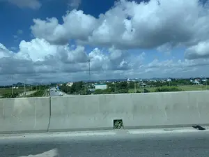 Bức ảnh được chụp từ trên một con đường trên cao hoặc cầu vượt, nhìn qua dải phân cách bằng bê tông. Phía dưới là một con đường có các phương tiện như ô tô và xe máy đang di chuyển. Xung quanh là cảnh quan vùng nông thôn với những cánh đồng xanh, cây cối và các khu dân cư. Một cột thu phát sóng viễn thông cao nổi bật trên nền trời. Ở phía xa, có thể nhìn thấy những dãy núi. Bầu trời trong xanh, có nhiều mây trắng. Có một số biển báo giao thông ở xa nhưng không thể đọc được nội dung.。（日本語訳）。写真は、高架道路または陸橋の上から、コンクリート製の中央分離帯越しに撮影されたものです。下には自動車やバイクなどの車両が走行している道路があります。周囲は緑の田畑、木々、住宅地のある田園風景です。高い通信用電波塔が空を背景に際立っています。遠方には、山々が見えます。空は青く澄み、白い雲が多く浮かんでいます。遠くにいくつかの交通標識がありますが、内容は読み取れません。