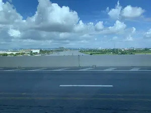 Bức ảnh được chụp từ trên một cây cầu hoặc đường cao tốc, nhìn ra một khung cảnh sông nước rộng lớn. Ở phía trước là mặt đường nhựa và một dải phân cách bằng bê tông. Phía sau dải phân cách là một con sông rộng, có thể là một cửa sông, với nhiều hoạt động của con người. Bờ bên trái của sông là một khu công nghiệp hoặc cảng biển với các nhà xưởng, cần cẩu lớn màu vàng, và các bồn chứa công nghiệp màu trắng. Nhiều tàu thuyền và sà lan đang neo đậu dọc bờ. Bờ bên phải có nhiều cây xanh, xen kẽ là các khu dân cư và các tòa nhà của một đô thị ở phía xa. Một vài chiếc thuyền đang di chuyển trên mặt sông. Bầu trời trong xanh và có nhiều mây trắng.。（日本語訳）。写真は橋または高速道路の上から撮影されたもので、広大な川の風景を見渡している。手前にはアスファルトの路面とコンクリートの中央分離帯がある。中央分離帯の後ろには広い川があり、それは河口かもしれず、多くの人々の活動が見られる。川の左岸は工業地帯または港で、工場、大きな黄色いクレーン、そして白い工業用タンクがある。多くの船やはしけが岸に沿って停泊している。右岸には緑の木々が多く、その間に住宅地や遠くの都市の建物が点在している。数隻のボートが川面を移動している。空は青く澄んでおり、白い雲が多く浮かんでいる。