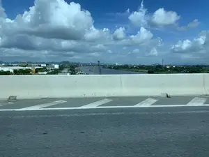 Bức ảnh được chụp từ trên một cây cầu hoặc đường cao tốc, nhìn ra một con sông lớn. Ở phía trước là mặt đường nhựa và dải phân cách bằng bê tông. Phía bên kia dải phân cách là một khu vực công nghiệp ven sông. Có nhiều cần cẩu (cần trục), cho thấy đây có thể là một cảng sông, một công trường xây dựng, hoặc một xưởng đóng tàu. Nhiều tàu thuyền và sà lan đang di chuyển hoặc neo đậu trên sông, phục vụ cho hoạt động vận tải. Dọc bờ sông có các nhà xưởng, kho bãi. Trên một tòa nhà màu trắng có thể đọc được một phần chữ "... NINH BÌNH". Ở phía xa là khung cảnh của một thành phố với các tòa nhà và những dãy núi trập trùng. Bầu trời trong xanh, có nhiều mây trắng.。（日本語訳）。写真は橋または高速道路の上から撮影されたもので、大きな川を見下ろしています。手前にはアスファルトの路面とコンクリートの中央分離帯があります。中央分離帯の向こう側は川沿いの工業地帯です。多くのクレーン（起重機）があり、ここが河川港、建設現場、または造船所である可能性を示しています。多くの船やはしけが川の上を移動または停泊しており、輸送活動に従事しています。川岸に沿って工場や倉庫があります。白い建物の上には「... NINH BÌNH」という文字の一部が読み取れます。遠くには建物や起伏のある山々が連なる街の風景が広がっています。空は青く澄んでおり、白い雲が多く浮かんでいます。