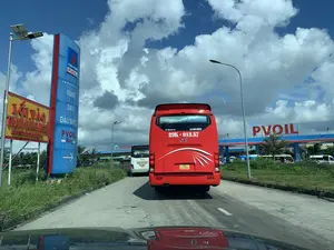 Bức ảnh chụp cảnh trên một con đường, nhìn từ trong một chiếc ô tô. Phía trước là một chiếc xe khách màu đỏ đang di chuyển về phía một trạm xăng và trạm dừng nghỉ. Các phương tiện dường như đang xếp hàng để vào trạm.。Các dòng chữ có thể đọc được bao gồm:。- Trên biển hiệu lớn màu xanh của trạm xăng: "PETROVIETNAM PVOIL", "XĂNG", "DẦU", "DẦU NHỚT", "PVOIL Easy" và một mũi tên chỉ lối vào.。- Trên biển báo màu vàng: "LỐI VÀO TRẠM DỪNG NGHỈ" cùng với mũi tên chỉ sang phải.。- Phía sau chiếc xe khách màu đỏ: "XE HỢP ĐỒNG" và "VP: 0905 673 119".。- Trên nóc trạm xăng phía xa: logo "PVOIL".。- Trên một chiếc xe khách màu trắng khác: logo "THACO".。Hoạt động chính là các phương tiện giao thông, chủ yếu là xe khách, đang tiến vào khu vực trạm xăng và trạm dừng nghỉ. Khung cảnh cho thấy một ngày nắng đẹp với bầu trời xanh và mây trắng. Ven đường có cỏ và hoa dại mọc xanh tốt. Có thể thấy các cơ sở vật chất như cột đèn đường, trong đó có một cột đèn năng lượng mặt trời.。（日本語訳）。ある道路の風景を車の中から撮影した写真です。前方には、ガソリンスタンドと休憩所に向かって移動している赤いバスがあります。車両は施設に入るために列を作っているようです。読み取れる文字は以下の通りです。- ガソリンスタンドの大きな青い看板には、「PETROVIETNAM PVOIL」、「XĂNG（ガソリン）」、「DẦU（軽油）」、「DẦU NHỚT（潤滑油）」、「PVOIL Easy」と入口を示す矢印があります。- 黄色い看板には、「LỐI VÀO TRẠM DỪNG NGHỈ（休憩所入口）」と右を指す矢印があります。- 赤いバスの後部には、「XE HỢP ĐỒNG（契約バス）」と「VP: 0905 673 119」と書かれています。- 遠くのガソリンスタンドの屋根には、「PVOIL」のロゴがあります。- 別の白いバスには、「THACO」のロゴがあります。主な動きは、主にバスなどの交通車両がガソリンスタンドと休憩所のエリアに入っていく様子です。風景は、青い空と白い雲のある晴れた美しい日であることを示しています。道端には草や野花が青々と生い茂っています。街灯などのインフラが見られ、その中にはソーラーパネル付きの街灯も1本あります。