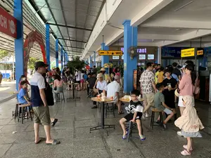 Bối cảnh là một không gian rộng lớn, đông đúc tại một trạm dừng nghỉ trên đường cao tốc. Nơi này có mái che cao bằng kim loại, được chống đỡ bởi các cột trụ sơn màu xanh. Rất nhiều người, bao gồm các gia đình có cả trẻ em và người lớn tuổi, đang nghỉ ngơi trong chuyến đi. Một số người đang ngồi tại các bàn tròn nhỏ để uống nước và trò chuyện, trong khi những người khác đang đứng xếp hàng tại khu vực quầy hàng. Các hoạt động chính là nghỉ ngơi, ăn uống và mua sắm. Có thể thấy các đồ vật như bàn ghế, quạt treo tường, cây cảnh trang trí, và một chiếc xe khách ở phía xa. Một người đàn ông trong ảnh đội nón lá.。Các dòng chữ và biển hiệu có thể đọc được:。- Trên cổng chào màu đỏ có logo Coca-Cola: "TRẠM DỪNG NGHỈ CAO TỐC... KÍNH CHÀO QUÝ KHÁCH".。- Biển hiệu màu vàng phía trên quầy hàng: "GỌI ĐỒ & THANH TOÁN" và "ORDER & PAY".。- Biển chỉ dẫn màu vàng treo trên cao có các biểu tượng mũi tên, dao dĩa, tách cà phê và chữ "WC", chỉ đường đến khu vực ăn uống và nhà vệ sinh.。- Tên "Restaurant" được chiếu sáng trên một quầy hàng.。- Biển hiệu "Cozy TAKE AWAY".。- Biển quảng cáo của Pepsi với khẩu hiệu "NGON QUÁ PEPSI ƠI。".。- Một biển hiệu khác có chữ "QUẦY CAFE TRÀ SỮA".。（日本語訳）。背景は、高速道路のサービスエリアにある、広くて混雑した空間です。この場所には金属製の高い屋根があり、青く塗られた柱で支えられています。子供からお年寄りまでいる家族連れを含む多くの人々が、移動の途中で休憩しています。何人かは小さな円卓に座って飲み物を飲んだりおしゃべりをしたりしており、他の人々は売店のエリアで列に並んでいます。主な活動は、休憩、飲食、買い物です。テーブルや椅子、壁掛け扇風機、装飾用の観葉植物、そして遠くには一台のバスが見えます。写真の中の一人の男性はノンラーをかぶっています。読み取れる文字や看板：。- コカ・コーラのロゴがある赤いウェルカムゲート：「高速道路サービスエリア…お客様を歓迎いたします」。- 売店の上にある黄色い看板：「ご注文とお支払い」と「ORDER & PAY」。- 高い位置に吊るされた黄色い案内板には、矢印、ナイフとフォーク、コーヒーカップのアイコンと「WC」の文字があり、飲食エリアとトイレへの道を示しています。- ある売店の上で「Restaurant」という名前がライトアップされています。- 「Cozy TAKE AWAY」という看板。- 「おいしいね、ペプシ。」というキャッチコピーのペプシの広告看板。- 「カフェ・ミルクティーカウンター」という文字がある別の看板。