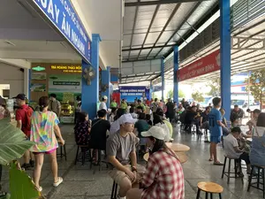 Các dòng chữ có thể đọc được trên các biển hiệu:。- Biển hiệu màu xanh dương: "...QUẦY ĂN NHANH" và logo Pepsi.。- Biển hiệu màu đỏ: "TRẠM DỪNG NGHỈ CAO TỐC..." và logo Coca-Cola.。- Biển hiệu tại quầy hàng: "TRÀ HOÀNG THẢO MỘC VÌ SỨC KHỎE AN KHANG" và "TRÀ CHO SỨC KHỎE BÌNH AN".。Đây là hình ảnh tại một trạm dừng nghỉ đông đúc ven đường cao tốc. Không gian của trạm được thiết kế mở, có mái che cao bằng kim loại. Rất đông người, bao gồm cả người lớn và trẻ em, có vẻ là hành khách, đang nghỉ ngơi. Họ ngồi trên những chiếc ghế đẩu thấp bằng gỗ và kim loại, quây quần bên những chiếc bàn tròn nhỏ để ăn uống và trò chuyện. Trong ảnh có một quầy hàng bán trà thảo mộc. Các vật thể khác có thể thấy là quạt treo tường, và ở phía sau là bãi đỗ xe có xe khách và ô tô.。（日本語訳）。看板で読み取れる文字：。- 青い看板：「...ファストフードカウンター」とペプシのロゴ。- 赤い看板：「高速道路サービスエリア...」とコカ・コーラのロゴ。- 売店の看板：「健康と安泰のためのロイヤルハーブティー」と「平穏な健康のためのお茶」。これは高速道路沿いの混雑したサービスエリアでの画像です。サービスエリアの空間はオープンな設計で、高い金属製の屋根があります。大人や子供を含む多くの人々、見たところ乗客のようですが、休憩しています。彼らは木と金属でできた低いスツールに座り、小さな円卓を囲んで飲食したりおしゃべりしたりしています。写真にはハーブティーを売る売店があります。