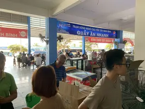 Bối cảnh là một trạm dừng nghỉ ven đường cao tốc đông đúc. Không gian bên trong là khu vực ăn uống và bán hàng không gian mở.。Chữ viết trên các biển hiệu:。- Biển hiệu chính lớn màu xanh dương có dòng chữ trắng: "TRẠM DỪNG NGHỈ CAO TỐC TÂY NINH BÌNH" và bên dưới là "QUẦY ĂN NHANH". Trên biển hiệu này còn có quảng cáo của Pepsi với dòng chữ "ĐÃAAAA QUÁ PEPSI NGON ƠI。".。- Ngay dưới biển hiệu màu xanh là một biển hiệu màu đỏ với chữ trắng: "KÍNH CHÀO QUÝ KHÁCH".。- Phía bên trái, một phần của biển hiệu màu đỏ có logo Coca-Cola và chữ "...H BÌNH".。- Ở phía sau, có một cổng chào màu đỏ cũng ghi "TRẠM DỪNG NGHỈ CAO TỐC TÂY NINH BÌNH".。- Trên một tủ đông có nhãn hiệu kem "Celano". Các thùng hàng có chữ "Dasani" và "QQ MINI CAKE".。Hoạt động và cảnh vật:。- Đây là một khu vực ăn uống và nghỉ ngơi. Nhiều người, có vẻ là hành khách, đang ngồi tại các bộ bàn ghế để ăn uống và nghỉ ngơi trong lúc chờ xe.。- Một số người khác đang đứng gần quầy hàng và các tủ đông, có thể đang mua đồ ăn, thức uống.。- Bên ngoài sân, nhiều xe khách lớn đang đỗ, cho thấy đây là điểm dừng chân của các tuyến xe đường dài.。- Không gian được trang bị nhiều quạt điện (quạt treo tường và quạt cây) để làm mát. Có các tủ kính trưng bày thực phẩm, tủ hấp và tủ đông lạnh để phục vụ khách hàng.。（日本語訳）。背景は、混雑した高速道路沿いのサービスエリアです。内部の空間は、オープンスペースの飲食・販売エリアです。看板の文字：。- 青地に白文字の大きなメイン看板：「TAY NINH BINH高速道路サービスエリア」、その下には「ファストフードカウンター」。この看板にはペプシの広告もあり、「ĐÃAAAA QUÁ PEPSI NGON ƠI。」（ああ、おいしいペプシ。）というキャッチコピーが書かれています。- 青い看板のすぐ下には、赤地に白文字の看板：「お客様を歓迎します」。- 左側には、コカ・コーラのロゴと「...H BINH」という文字が入った赤い看板の一部が見えます。- 後方には、「TAY NINH BINH高速道路サービスエリア」と書かれた赤いウェルカムゲートもあります。- 冷凍庫には「Celano」というアイスクリームのブランド名があります。段ボール箱には「Dasani」と「QQ MINI CAKE」の文字が見えます。活動と風景：。- ここは飲食と休憩のエリアです。多くの人々、おそらく乗客が、テーブルと椅子のセットに座って食事をしたり、バスを待つ間に休憩したりしています。- 他の人々は、売店や冷凍庫の近くに立っており、食べ物や飲み物を買っているようです。- 外の駐車場には多くの大型バスが停車しており、ここが長距離バス路線の休憩地点であることを示しています。- 空間には涼を取るための多くの扇風機（壁掛け扇風機とスタンド扇風機）が設置されています。食品を陳列するガラスケース、蒸し器、冷凍庫があり、顧客にサービスを提供しています。