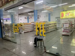 Bối cảnh là bên trong một cửa hàng tiện lợi hoặc cửa hàng đặc sản tại một trạm dừng nghỉ, được chụp từ bên ngoài qua cửa kính. Một người đàn ông, có thể là nhân viên, đang ngồi trên một chiếc ghế đẩu gần cửa ra vào. Bên trong cửa hàng, các kệ hàng được sắp xếp để trưng bày nhiều loại sản phẩm như đồ ăn nhẹ và đặc sản đóng gói, mặc dù một số kệ hàng còn trống. Sàn nhà được lát gạch, và trên trần nhà có đèn huỳnh quang và quạt trần. Một bình chữa cháy được đặt cạnh cửa.。Các văn bản được ghi lại:。- Trên cửa kính: 'COFFEE', 'SPECIAL BREW', 'OPEN 08:00', 'CLOSE 21:00', 'ICE TEA & COFFEE ARE OUT'.。- Biển hiệu lớn bên trong: 'CÔNG TY CP SẢN XUẤT THƯƠNG MẠI ĐẠI LONG', 'OCOP', 'TRẠM DỪNG NGHỈ TÂY NINH BÌNH'.。- Các biển hiệu nhỏ hơn: 'NƯỚC GIẢI KHÁT', 'NEM CÁC LOẠI', 'Cơm cháy'.。（日本語訳）。背景は、サービスエリアにあるコンビニエンスストアまたは特産品店の中であり、ガラスのドア越しに外から撮影されています。おそらく従業員と思われる男性が、出入り口の近くのスツールに座っています。店内では、スナック菓子やパッケージされた特産品など、さまざまな種類の商品を陳列するために商品棚が並べられていますが、いくつかの棚は空です。床はタイル張りで、天井には蛍光灯とシーリングファンがあります。ドアの横に消火器が置かれています。記録されているテキスト：。- ガラスのドアに：「COFFEE」、「SPECIAL BREW」、「OPEN 08:00」、「CLOSE 21:00」、「ICE TEA & COFFEE ARE OUT」。- 店内の大きな看板：「CÔNG TY CP SẢN XUẤT THƯƠNG MẠI ĐẠI LONG」、「OCOP」、「TRẠM DỪNG NGHỈ TÂY NINH BÌNH」。- 小さな看板：「NƯỚC GIẢI KHÁT」、「NEM CÁC LOẠI」、「Cơm cháy」。