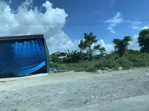Hình ảnh chụp một khung cảnh ven đường ở khu vực nông thôn. Phía trước là một con đường đất đá. Bên trái có một công trình giống như nhà kho hoặc lán trại tạm, có mái tôn và được che bằng một tấm bạt lớn màu xanh dương. Ở ven đường có một ít rác thải và cỏ dại mọc um tùm. Phía sau là những ngôi nhà dân, cây cối xanh tươi, trong đó có thể thấy cả cây chuối. Bầu trời trong xanh, có nhiều mây trắng và có đường dây điện chạy ngang qua. Không có hoạt động nào của con người được ghi nhận.。（日本語訳）。田舎の道端の風景を撮影した写真です。手前には砂利道があります。左側には、倉庫か仮設の小屋のような建物があり、トタン屋根で、大きな青いシートで覆われています。道端には少量のゴミがあり、雑草が生い茂っています。後方には民家や青々とした木々があり、その中にはバナナの木も見えます。空は青く澄んでおり、白い雲が多く、電線が横切っています。人の活動は見られません。