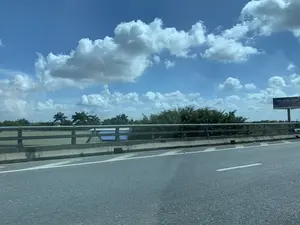 Bức ảnh được chụp từ một phương tiện đang di chuyển trên đường cao tốc hoặc cầu vượt vào một ngày nắng đẹp. Bầu trời trong xanh với nhiều đám mây trắng. Bên phải đường có một biển quảng cáo lớn ghi: "THÉP KYOEI JAPAN", bên dưới là dòng chữ "Cùng uy tín - Chất lượng Nhật" và một dòng chữ tiếng Nhật "日本の鉄は日本の品質" cùng với lá cờ Nhật Bản. Nhìn qua lan can cầu, có thể thấy cây cối xanh tươi, một vài cây cọ và mái nhà ở phía xa. Phần tiền cảnh là mặt đường nhựa và dải phân cách bê tông của cây cầu.。（日本語訳）。この写真は、晴れた美しい日に、高速道路または高架橋を走行中の乗り物から撮影されたものです。空は青く澄み渡り、白い雲が多く浮かんでいます。道路の右側には、「THÉP KYOEI JAPAN」と書かれた大きな広告看板があり、その下には「Cùng uy tín - Chất lượng Nhật」という文言と、「日本の鉄は日本の品質」という日本語の文言が日本の国旗と共に記されています。橋の欄干越しには、青々とした木々、数本のヤシの木、そして遠くには家々の屋根が見えます。