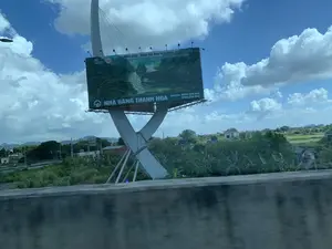 Bức ảnh chụp một tấm biển quảng cáo lớn bên đường, có thể là trên một tuyến đường cao tốc, vào một ngày trời xanh có mây. Tấm biển được đỡ bởi một kết cấu cột trụ lớn màu trắng có thiết kế hiện đại. Phía sau tấm biển là khung cảnh nông thôn với cây cối xanh tươi, một vài ngôi nhà, và những ngọn núi ở phía xa. Ở tiền cảnh có một lan can bê tông của cầu hoặc đường trên cao, cho thấy ảnh được chụp từ một phương tiện đang di chuyển. Dưới chân biển quảng cáo có một con đường nhỏ với vài người đang đi xe máy.。Nội dung chữ viết trên biển quảng cáo:。- Phía trên: "NINH BÌNH - Điểm đến thân thiện hàng đầu thế giới。Ninh Bình - The top ten friendliest destinations in the World。". Hình ảnh trên biển quảng cáo là một cảnh quan sông nước với nhiều thuyền du lịch, đặc trưng của tỉnh Ninh Bình.。- Phía dưới: "NHÀ HÀNG THANH NGA" và thông tin liên hệ "Hotline: 0858.228.868, 0858.280.092".。（日本語訳）。写真は、雲のある青空の日に、おそらく高速道路上の道端にある大きな広告看板を写したものである。看板は、モダンなデザインの大きな白い支柱構造によって支えられている。看板の後ろには、青々とした木々、いくつかの家、そして遠くの山々といった田園風景が広がっている。前景には橋または高架道路のコンクリート製の手すりがあり、写真が移動中の乗り物から撮影されたことを示している。広告看板の下には小さな道があり、数人がバイクに乗っている。広告看板に書かれている内容：。- 上部：「ニンビン - 世界で最もフレンドリーな目的地。ニンビン - 世界で最もフレンドリーな目的地トップ10。」。広告看板の写真は、多くの観光船が浮かぶ水郷の風景であり、ニンビン省の特徴である。- 下部：「タイン・ガー・レストラン」と連絡先情報「ホットライン：0858.228.868、0858.280.092」。