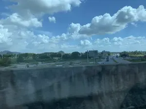 Bức ảnh được chụp từ một phương tiện đang di chuyển, nhìn ra khung cảnh bên ngoài qua cửa sổ. Khung cảnh là một con đường quốc lộ hoặc cao tốc chạy qua vùng nông thôn. Bầu trời trong xanh với nhiều đám mây trắng. Ở phía xa là những dãy núi đá vôi và cây cối xanh tươi. Trên đường có các phương tiện giao thông như xe tải, ô tô và xe máy đang di chuyển. Có một số biển báo giao thông lớn màu xanh lá cây nhưng không thể đọc rõ chữ. Ở tiền cảnh, một phần lan can cầu hoặc thành xe bị mờ. Bên đường có các khu đất với cây cối, một ao nước nhỏ, và các đống vật liệu trông giống như cát hoặc sỏi, cho thấy có thể có hoạt động xây dựng gần đó.。（日本語訳）。移動中の乗り物から窓越しに外の景色を撮影した写真です。景色は、田園地帯を走る国道または高速道路です。空は青く澄んでおり、白い雲がたくさん浮かんでいます。遠くには石灰岩の山々と青々とした木々があります。道路上では、トラック、乗用車、バイクなどの乗り物が走行しています。いくつかの大きな緑色の交通標識がありますが、文字ははっきりと読めません。