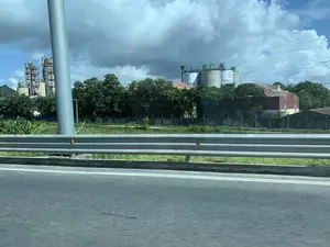 Trên các silo lớn màu trắng của nhà máy có ghi dòng chữ 'XI MĂNG DUYÊN HÀ' cùng với logo. Khung cảnh được chụp từ một phương tiện đang di chuyển trên đường, có thể là đường cao tốc, với lan can bảo vệ ở phía trước. Nhìn qua lan can là một khu công nghiệp, nổi bật là một nhà máy xi măng lớn. Nhà máy bao gồm các tháp sản xuất cao và các silo chứa thành phẩm. Giữa đường và nhà máy là một khu đất trồng cây, bao gồm cả cây chuối, được bao quanh bởi một bức tường bê tông. Bầu trời nhiều mây, và có các đường dây điện chạy ngang qua.。（日本語訳）。工場の大きな白いサイロには、「XI MĂNG DUYÊN HÀ」という文字とロゴが書かれています。この風景は、道路を移動中の乗り物から撮影されたもので、おそらく高速道路であり、手前にはガードレールがあります。ガードレールの向こうには工業地帯が見え、大きなセメント工場が目立っています。工場は、高い生産塔と完成品を貯蔵するサイロで構成されています。道路と工場の間には、バナナの木を含む木々が植えられた土地があり、コンクリートの壁で囲まれています。空は曇っており、電線が横切っています。