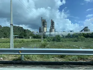 Khung cảnh cho thấy một nhà máy công nghiệp lớn, rất có thể là nhà máy xi măng, nằm giữa vùng nông thôn. Nhà máy có các cấu trúc đặc trưng như silo chứa liệu, các tháp lò cao và hệ thống băng tải. Phía sau nhà máy là những ngọn núi đá vôi xanh rì. Phía trước là một khu đất trống với cây cỏ và các vũng nước, trông giống như một cánh đồng bị ngập hoặc một vùng đầm lầy. Bức ảnh được chụp từ một phương tiện đang di chuyển trên đường, có thể thấy lan can đường ở phía trước. Bầu trời phía trên có cả mây trắng và những đám mây xám dày đặc. Có nhiều cột điện và đường dây điện trong khu vực. Không có văn bản hay biển hiệu nào có thể đọc được.。（日本語訳）。風景は、田園地帯の中に位置する大きな工業工場、おそらくセメント工場を示しています。工場には、原料貯蔵サイロ、高炉塔、ベルトコンベアシステムといった特徴的な構造物があります。工場の後ろには、青々とした石灰岩の山々があります。前方には草木と水たまりのある空き地があり、浸水した野原か湿地帯のように見えます。写真は道路を移動中の乗り物から撮影されたもので、手前には道路のガードレールが見えます。上空には白い雲と厚い灰色の雲の両方があります。この地域には多くの電柱と電線があります。読み取れるテキストや標識はありません。