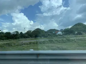 Không có thông tin văn bản nào có thể đọc được. Hình ảnh cho thấy một phong cảnh nông thôn, được chụp từ một phương tiện đang di chuyển trên đường. Ở tiền cảnh là một dải hộ lan bằng kim loại. Phía sau hộ lan là một khu đất trống với cỏ dại. Xa hơn là những ngôi nhà dân có mái ngói nằm xen kẽ giữa cây cối xanh tươi. Phía sau là những ngọn đồi được bao phủ bởi cây xanh. Bầu trời trong xanh và có nhiều mây trắng. Không có hoạt động cụ thể nào của con người được ghi lại.。（日本語訳）。読み取れるテキスト情報はありません。画像は、道路を移動中の乗り物から撮影された、田舎の風景を示しています。前景には金属製のガードレールがあります。ガードレールの後ろには雑草の生えた空き地があります。その向こうには、緑豊かな木々の間に瓦屋根の民家が点在しています。その後ろには緑に覆われた丘があります。空は青く、白い雲が多く浮かんでいます。特定の人間活動は記録されていません。