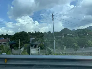 Bức ảnh chụp một khung cảnh nông thôn ở Việt Nam, có thể được chụp từ một phương tiện đang di chuyển do có lan can đường ở phía trước. Cảnh vật bao gồm nhiều ngôi nhà dân nằm rải rác giữa cây cối xanh tươi, với một số cây chuối có thể nhận ra. Một cột điện bê tông và các đường dây điện chạy ngang qua. Ở phía sau, những ngọn đồi và núi đá vôi được bao phủ bởi thảm thực vật dày đặc nổi bật trên nền trời xanh có nhiều mây. Không có hoạt động cụ thể nào của con người được quan sát, chỉ có một cảnh quan tĩnh lặng và yên bình. Không có văn bản hay biển hiệu nào có thể đọc được.。（日本語訳）。この写真はベトナムの田舎の風景を写したもので、手前に道路のガードレールがあることから、移動中の乗り物から撮影された可能性があります。風景には、青々とした木々の間に点在する多くの民家が含まれており、いくつかのバナナの木が見分けられます。コンクリートの電柱と電線が横切っています。背景には、密集した植生に覆われた丘や石灰岩の山々が、雲の多い青空を背景に際立っています。特定の人の活動は見られず、静かで平和な景観だけがあります。読み取れる文字や看板はありません。