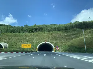 Bức ảnh chụp lối vào một đường hầm xuyên núi, nhìn từ trong một chiếc ô tô đang di chuyển. Con đường nhựa có nhiều làn được phân chia rõ ràng bằng vạch kẻ đường và các mũi tên chỉ hướng đi thẳng vào hầm. Phía trên và xung quanh cửa hầm là sườn đồi được gia cố và phủ đầy cỏ cùng cây xanh. Bầu trời trong xanh, có ít mây trắng. Có nhiều biển báo giao thông được đặt trước cửa hầm. Một biển báo lớn màu vàng bên trái có dòng chữ 'BẬT ĐÈN PHA' và 'TURN ON HEADLIGHT'. Phía trên cửa hầm có biển báo hình tròn giới hạn chiều cao '5.0m'. Bên phải lối vào có một cụm biển báo khác, bao gồm biển cấm dừng và đỗ xe, biển yêu cầu giữ khoảng cách tối thiểu 100m, và biển cấm sử dụng còi. Bên trái cửa hầm chính có một cửa hầm phụ nhỏ hơn.。（日本語訳）。移動中の車内から見た、山を貫くトンネルの入口を撮影した写真です。アスファルトの道路には複数の車線があり、車線とトンネルにまっすぐ向かう方向を示す矢印によって明確に区切られています。トンネルの入口の上と周りは、補強され草木で覆われた丘の斜面です。空は青く澄んでおり、白い雲が少しあります。トンネルの入口の前には多くの交通標識が設置されています。左側にある大きな黄色の標識には、「BẬT ĐÈN PHA」と「TURN ON HEADLIGHT」という文字があります。トンネルの入口の上には、高さ制限「5.0m」の円形標識があります。入口の右側には、駐停車禁止の標識、最低100mの車間距離を保つよう求める標識、そして警笛使用禁止の標識を含む、別の標識群があります。主トンネルの入口の左側には、より小さな副トンネルの入口があります。