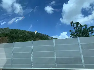 Bức ảnh được chụp từ bên trong một chiếc ô tô đang di chuyển, nhìn ra ngoài qua cửa sổ. Ở tiền cảnh là một hàng rào chắn màu xám, có thể là tấm chắn tiếng ồn của đường cao tốc. Phía sau hàng rào là một ngọn đồi được bao phủ bởi cây cối xanh tươi. Nổi bật trên đỉnh đồi là một bức tượng lớn màu trắng và vàng, có hình dạng một con rắn thần thoại với nhiều đầu, có thể là tượng thần Naga. Bầu trời trong xanh với vài đám mây trắng. Trên cửa kính xe có hình ảnh phản chiếu của nội thất bên trong xe và bóng người.。（日本語訳）。写真は、移動中の車内から窓の外を見て撮影されたものです。前景には灰色の防護柵があり、おそらく高速道路の防音壁でしょう。柵の後ろには、青々とした木々に覆われた丘があります。丘の頂上には、白と金色の大きな像が際立っており、複数の頭を持つ神話上の蛇の形をしていて、おそらくナーガ神の像でしょう。空は青く澄んでおり、白い雲がいくつか浮かんでいます。車の窓ガラスには、車内の内装と人影が映り込んでいます。