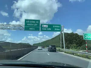 Bức ảnh chụp cảnh giao thông trên một tuyến đường cao tốc hoặc quốc lộ ở Việt Nam, được chụp từ bên trong một chiếc ô tô đang di chuyển. Bầu trời trong xanh, có nắng và nhiều mây trắng. Trên đường có một vài chiếc ô tô khác đang lưu thông. Phía trên có một giàn biển báo lớn màu xanh lá cây cung cấp thông tin cho người lái xe. Nội dung các biển báo như sau: Biển báo bên trái ghi 'DẢI DỪNG XE KHẨN CẤP' và '800 m'. Biển báo bên phải chỉ dẫn khoảng cách đến các địa điểm: 'QL.47 21.11 Km' và 'NGHI SƠN 31 Km'. Ngoài ra, ở phía xa bên phải có một biển báo cột mốc ghi số '200'. Con đường có nhiều làn xe, với dải phân cách cứng bằng bê tông và lan can bảo vệ. Phong cảnh hai bên đường là đồi núi trập trùng được bao phủ bởi cây cối xanh tươi.。（日本語訳）。ベトナムの高速道路または国道での交通風景を撮影した写真で、移動中の車内から撮影されたものです。空は澄み切った青で、晴れており、白い雲が多く浮かんでいます。道路上には、他にも数台の車が走行しています。上方には、ドライバーに情報を提供する大きな緑色の標識の骨組みがあります。標識の内容は以下の通りです。左側の標識には「DẢI DỪNG XE KHẨN CẤP」と「800 m」と書かれています。右側の標識は各地点までの距離を示しており、「QL.47 21.11 Km」と「NGHI SƠN 31 Km」とあります。さらに、右側の遠方には「200」という数字が書かれたキロポスト標識があります。道路は複数車線で、コンクリート製の中央分離帯とガードレールが設置されています。道路の両側の風景は、青々とした木々に覆われた起伏のある丘や山です。