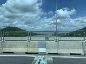 Bức ảnh được chụp từ một phương tiện đang di chuyển trên một cây cầu hoặc đường cao tốc. Cảnh vật được nhìn qua lan can của cây cầu.。- **Loại địa điểm:** Một cây cầu lớn bắc qua một con sông rộng.。- **Hoạt động:** Không có hoạt động cụ thể nào của con người được ghi lại, chỉ là cảnh quan nhìn từ một phương tiện đang chạy.。- **Các vật thể trong ảnh:**。- **Cấu trúc cầu:** Dải phân cách bằng bê tông, lan can kim loại, một cột đèn đường và mặt đường nhựa.。- **Phong cảnh:** Một con sông rộng lớn có nước màu nâu phù sa. Ở phía xa là những dãy đồi núi được bao phủ bởi cây xanh. Ven sông có một vài ngôi nhà và khu dân cư. Một cột điện cao thế có thể được nhìn thấy trên một ngọn đồi.。- **Bầu trời:** Bầu trời có màu xanh và nhiều mây trắng, xám.。- **Chữ viết:** Không có chữ viết nào có thể đọc được trong ảnh.。（日本語訳）。写真は、橋または高速道路を移動中の乗り物から撮影されたものです。景色は橋の欄干越しに見えます。- **場所の種類：** 幅広い川にかかる大きな橋。- **活動：** 特定の人間の活動は記録されておらず、走行中の乗り物から見た風景だけです。- **写真の中の物体：**。- **橋の構造物：** コンクリート製の中央分離帯、金属製の欄干、街灯柱1本、アスファルトの路面。- **風景：** 沖積土で茶色い水が流れる広大な川。遠くには緑に覆われた丘陵や山々が連なっています。川沿いにはいくつかの家や住宅地があります。丘の上には高圧電線の鉄塔が見えます。- **空：** 空は青く、白や灰色の雲が多く浮かんでいます。- **文字：** 写真には読み取れる文字はありません。