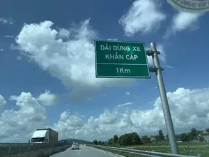 Bức ảnh chụp cảnh một con đường cao tốc vào ban ngày, dưới bầu trời xanh và nhiều mây trắng. Ở bên phải đường có một biển báo giao thông màu xanh lá cây với dòng chữ màu trắng: 'DẢI DỪNG XE KHẨN CẤP' và '1Km', báo hiệu rằng có một làn dừng xe khẩn cấp ở phía trước 1 km. Trên đường, có một chiếc ô tô con màu trắng và một xe tải container màu trắng đang di chuyển. Con đường có lan can bảo vệ. Nhìn sang hai bên đường là quang cảnh nông thôn với cây cối, đồng ruộng và nhà cửa.。（日本語訳）。青空と多くの白い雲の下、日中の高速道路の風景を撮った写真です。道路の右側には、「緊急停止帯」と「1Km」という白い文字が書かれた緑色の交通標識があり、1km先に緊急停止帯があることを示しています。道路上では、一台の白い乗用車と一台の白いコンテナトラックが走行しています。道路にはガードレールがあります。道路の両側を見ると、木々、田畑、家々のある田園風景が広がっています。