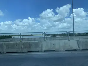 Bức ảnh được chụp từ một phương tiện đang di chuyển trên một cây cầu. Phía trước là dải phân cách bằng bê tông và lan can kim loại của cây cầu. Nhìn qua lan can là một con sông rộng. Ở phía xa bên kia sông là một khu dân cư với nhiều nhà cửa và cây cối. Bầu trời trong xanh và có nhiều mây trắng. Một cột đèn đường có thể được nhìn thấy ở phía bên phải.。（日本語訳）。写真は、橋の上を移動中の乗り物から撮影されたものです。前方には、コンクリート製の中央分離帯と橋の金属製の手すりがあります。手すりの向こうには広い川があります。川の向こうの遠くには、多くの家や木々がある住宅地があります。空は青く澄んでおり、白い雲がたくさんあります。右側には街灯が見えます。