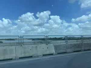 Bức ảnh được chụp từ một phương tiện đang di chuyển trên một cây cầu hoặc đường cao tốc. Ở tiền cảnh là dải phân cách bằng bê tông và lan can an toàn của cầu. Nhìn qua lan can là một vùng sông nước rộng lớn, có thể là một con sông hoặc hồ. Bên kia bờ là cảnh quan làng quê với những ngôi nhà, cây cối xanh tươi, và ở phía xa có thể thấy những ngọn đồi thấp. Bầu trời trong xanh và có nhiều mây trắng. Không có hoạt động cụ thể nào của con người được ghi lại, chỉ có cảnh quan nhìn từ trên cầu khi đang di chuyển.。（日本語訳）。写真は、橋または高速道路の上を移動中の乗り物から撮影されたものです。前景には、コンクリート製の中央分離帯と橋の安全欄干があります。欄干越しには広大な水域が見え、それは川か湖かもしれません。対岸には、家々や青々とした木々がある田舎の風景が広がり、遠くには低い丘が見えます。空は青く澄んでおり、白い雲がたくさん浮かんでいます。