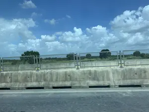 Bức ảnh được chụp từ một phương tiện đang di chuyển, có thể là trên một cây cầu hoặc đường cao tốc. Ở phía trước là một dải phân cách bằng bê tông và lan can kim loại. Nhìn qua lan can là khung cảnh vùng nông thôn với những cánh đồng nông nghiệp xanh mướt và cây cối rải rác. Bầu trời trong xanh, có nhiều đám mây trắng. Không có người hay hoạt động cụ thể nào được nhìn thấy trong các cánh đồng. Không có chữ viết hay biển báo nào có thể đọc được.。（日本語訳）。写真は移動中の乗り物から撮影されたもので、橋の上か高速道路の上である可能性があります。手前にはコンクリート製の中央分離帯と金属製の手すりがあります。手すりの向こうには、青々とした農地と点在する木々がある田園風景が広がっています。空は青く澄んでおり、白い雲が多く浮かんでいます。畑には人や特定の活動は見られません。読み取れる文字や標識はありません。