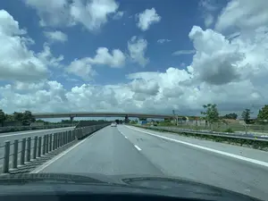 Bức ảnh ghi lại khung cảnh trên một tuyến đường cao tốc hiện đại, được chụp từ góc nhìn bên trong một chiếc ô tô đang di chuyển. Con đường có nhiều làn xe, mặt đường nhựa, với dải phân cách và lan can bảo vệ chắc chắn ở hai bên. Phía trước có một chiếc ô tô màu trắng đang chạy cùng chiều. Một cây cầu vượt bằng bê tông bắc ngang qua đường cao tốc. Bầu trời trong xanh, có nhiều mây trắng, cho thấy thời tiết đẹp. Hai bên đường là cây cối và hàng rào. Ở phía xa bên phải có một số biển báo giao thông, trong đó có một biển màu xanh lam nhưng chữ viết không thể đọc rõ hoàn toàn.。（日本語訳）。写真は、移動中の車内からの視点で撮影された、現代的な高速道路上の光景を捉えている。道路は複数の車線とアスファルトの路面を持ち、両側には中央分離帯と頑丈なガードレールがある。前方には、同じ方向に走っている白い車が一台いる。コンクリート製の陸橋が高速道路を横切って架かっている。空は青く澄み、白い雲が多く、天気が良いことを示している。道路の両側には木々とフェンスがある。右手の遠方にはいくつかの交通標識があり、その中には青い標識が一つあるが、文字は完全にはっきりと読むことはできない。