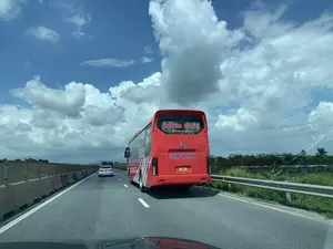 Bức ảnh được chụp từ bên trong một chiếc ô tô đang di chuyển trên đường cao tốc vào một ngày nắng đẹp. Phía trước là một chiếc xe khách lớn màu đỏ, trên kính sau có ghi tên nhà xe 'KHIÊM OANH' và dòng chữ 'Limousine', 'UNIVERSE'. Ở làn đường bên trái, một chiếc ô tô con màu trắng đang đi phía trước. Con đường có nhiều làn xe, được ngăn cách bằng dải phân cách bê tông và lan can kim loại. Hai bên đường là cây cối xanh tươi. Bầu trời trong xanh và có nhiều mây trắng lớn.。（日本語訳）。この写真は、晴れた美しい日に高速道路を走行中の車内から撮影されたものです。前方には大きな赤いバスがおり、後部ガラスにはバス会社の名前「KHIÊM OANH」と「Limousine」、「UNIVERSE」という文字が書かれています。左側の車線では、白い乗用車が前方を走っています。道路は複数車線あり、コンクリートの中央分離帯と金属製のガードレールで仕切られています。道路の両側には青々とした木々があります。空は青く澄んでおり、大きな白い雲がたくさん浮かんでいます。