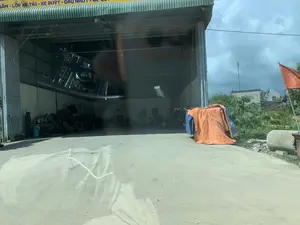 Bản ghi văn bản trên biển hiệu màu vàng chữ xanh: '...SĂM - LỐP XE TẢI - XE BUÝT - DẦU NHỚT - ẮC QUY...'. Bối cảnh là một gara hoặc xưởng sửa chữa ô tô lớn, có vẻ chuyên về xe tải và xe buýt. Xưởng có kết cấu khung thép, mái tôn, với cửa cuốn đang mở rộng. Bên trong xưởng, có thể thấy nhiều lốp xe cũ xếp chồng lên nhau, một máy nén khí và các bình khí nén. Phía trước là một khoảng sân bê tông. Ở bên phải, có một vật được che phủ bởi một tấm bạt màu cam và một lá cờ đỏ sao vàng được cắm gần đó. Bầu trời nhiều mây. Hình ảnh được chụp từ bên trong một chiếc xe, có hình ảnh phản chiếu của bảng điều khiển xe trên kính.。（日本語訳）。黄色の看板に青い文字で書かれたテキストの記録：「...チューブ - トラック用タイヤ - バス用タイヤ - エンジンオイル - バッテリー...」。背景は大きな自動車修理工場またはガレージで、トラックやバスを専門としているようです。工場は鉄骨構造で、トタン屋根を持ち、シャッターが大きく開いています。工場の中には、積み重ねられた多くの古いタイヤ、エアコンプレッサー、そして圧縮ガスボンベが見えます。正面にはコンクリートの広場があります。右側には、オレンジ色のシートで覆われた物体があり、その近くに金星紅旗が立てられています。空は曇っています。写真は車内から撮影されたもので、ガラスには車のダッシュボードの反射が映っています。