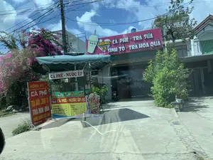 Đây là hình ảnh mặt tiền của một quán nước giải khát tên là 'Hồng Vy', nằm ven một con đường bê tông. Quán có nhiều biển hiệu quảng cáo các loại đồ uống. Biển hiệu chính lớn màu hồng có ghi 'HỒNG VY', 'CÀ PHÊ - TRÀ SỮA', 'SINH TỐ - TRÀ HOA QUẢ'. Phía trước quán có một xe đẩy bán hàng di động với mái che, trên xe có biển hiệu 'CÀ PHÊ - NƯỚC ÉP' và một bảng thực đơn liệt kê các loại đồ uống như: Cà Phê Nâu, Cà Phê Đen, Cà Phê Muối, Cà Phê Cốt Dừa, và các loại nước ép như Ép Cam, Ép Táo, Ép Dưa Hấu, Ép Dứa, Ép Ổi, Ép Cóc. Ngoài ra còn có một biển hiệu đứng màu đỏ ghi 'HỒNG VY', 'CÀ PHÊ', 'TRÀ SỮA - SINH TỐ', 'TRÀ HOA QUẢ', 'NƯỚC UỐNG CÁC LOẠI'. Trên cửa kính của quán có chữ 'coffee | Trà sữa'. Khung cảnh xung quanh có cây xanh và một giàn hoa giấy màu hồng rực rỡ. Phía trên là bầu trời xanh có mây trắng và nhiều dây điện chằng chịt trên cột điện. Một chiếc xe máy đang đậu bên trong hiên quán.。（日本語訳）。これは「Hồng Vy」という名前の、コンクリートの道端にある飲料店の正面の写真です。店には様々な種類の飲み物を宣伝する多くの看板があります。ピンク色の大きなメイン看板には「HỒNG VY」、「CÀ PHÊ - TRÀ SỮA」（コーヒー - ミルクティー）、「SINH TỐ - TRÀ HOA QUẢ」（スムージー - フルーツティー）と書かれています。店の前には屋根付きの移動販売カートがあり、カートには「CÀ PHÊ - NƯỚC ÉP」（コーヒー - ジュース）という看板と、Cà Phê Nâu（ブラウンコーヒー）、Cà Phê Đen（ブラックコーヒー）、Cà Phê Muối（塩コーヒー）、Cà Phê Cốt Dừa（ココナッツコーヒー）などのコーヒー類、Ép Cam（オレンジジュース）、Ép Táo（リンゴジュース）、Ép Dưa Hấu（スイカジュース）、Ép Dứa（パイナップルジュース）、Ép Ổi（グアバジュース）、Ép Cóc（アンバレラジュース）などのジュース類がリストされたメニューボードがあります。その他にも、「HỒNG VY」、「CÀ PHÊ」（コーヒー）、「TRÀ SỮA - SINH TỐ」（ミルクティー - スムージー）、「TRÀ HOA QUẢ」（フルーツティー）、「NƯỚC UỐNG CÁC LOẠI」（各種飲料）と書かれた赤い立て看板があります。店のガラス戸には「coffee | Trà sữa」（コーヒー | ミルクティー）という文字があります。周囲の風景には緑の木々と、鮮やかなピンク色のブーゲンビリアの棚があります。上には白い雲のある青空と、電柱に張り巡らされた多くの電線があります。一台のバイクが店の軒下に停まっています。