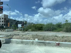 Bức ảnh chụp một khu vực đang trong quá trình xây dựng, có thể là một khu đô thị mới hoặc một ngã tư đường. Ở phía sau là một tòa nhà nhiều tầng đang được xây dựng dở dang. Cảnh quan xung quanh còn khá hoang sơ với đất đá, các khối bê tông và cỏ dại mọc um tùm. Có một cột đèn tín hiệu giao thông đang bật đèn đỏ và đồng hồ đếm ngược hiển thị số '04'. Một băng rôn màu đỏ được treo lên với nội dung: '2024', 'MỪNG ĐẤT NƯỚC ĐỔI MỚI', 'MỪNG ĐẢNG QUANG VINH'. Ngoài ra còn có một tấm biển thông tin dự án lớn màu trắng với sơ đồ quy hoạch, nhưng nội dung chi tiết không thể đọc được. Bầu trời trong xanh, có nhiều mây trắng.。（日本語訳）。写真は建設中の地域を写しており、新しい都市区か道路の交差点かもしれません。後方には、建設途中の多層階の建物があります。周囲の景観はまだかなり荒れており、土や石、コンクリートブロック、そして生い茂る雑草があります。交通信号機の柱が1本あり、赤信号が点灯していて、カウントダウンタイマーには「04」という数字が表示されています。赤い横断幕が掲げられており、その内容は「2024」、「国の刷新を祝う」、「栄光ある党を祝う」です。さらに、計画図が描かれた大きな白いプロジェクト情報看板もありますが、詳細な内容は読み取れません。空は青く澄んでおり、白い雲が多く浮かんでいます。