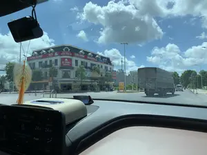 Bức ảnh được chụp từ bên trong một chiếc ô tô, nhìn ra một ngã tư hoặc vòng xuyến trên đường phố vào một ngày nắng. Ở phía bên trái là một tòa nhà lớn, nhiều tầng với các biển hiệu có thể đọc được như: 'GLOBAL ACADEMY', 'IELTS', 'GLOBAL DONG SON', 'ENGLISH COFFEE', 'VILAZA TRUNG GIANG'. Dưới tầng trệt có các biển hiệu khác như 'CƠM PHÚ BÌNH DÂN - XE CON - XE TẢI' và một biển hiệu màu vàng ghi 'PHỞ TRÂU BÚN CÁ CƠM BÌNH DÂN'. Hoạt động chính là giao thông trên đường, với một chiếc xe tải lớn có mui bạt đang di chuyển phía trước, và các phương tiện khác ở phía xa. Bên trong xe, có thể thấy bảng điều khiển, một camera hành trình, và một vật trang trí hình lá bồ đề treo trên gương chiếu hậu. Màn hình trên xe hiển thị thời gian là 9:58 và nhiệt độ là 24.0°C. Bầu trời trong xanh và có nhiều mây.。（日本語訳）。写真は晴れた日に、車内から街路の交差点またはロータリーを眺めて撮影されたものです。左側には大きな多階建ての建物があり、「GLOBAL ACADEMY」、「IELTS」、「GLOBAL DONG SON」、「ENGLISH COFFEE」、「VILAZA TRUNG GIANG」といった読み取れる看板があります。1階には「CƠM PHÚ BÌNH DÂN - XE CON - XE TẢI」のような他の看板や、「PHỞ TRÂU BÚN CÁ CƠM BÌNH DÂN」と書かれた黄色い看板があります。主な活動は道路上の交通で、前方を幌付きの大型トラックが走行しており、遠くには他の車両も見えます。車内には、ダッシュボード、ドライブレコーダー、そしてバックミラーに吊るされた菩提樹の葉の形の装飾品が見えます。車内のディスプレイには、時刻が9時58分、気温が24.0℃と表示されています。空は青く澄んでおり、雲が多く浮かんでいます。