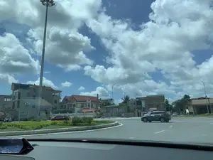 Bức ảnh chụp cảnh quan một vòng xuyến giao thông (bùng binh) từ góc nhìn bên trong một chiếc ô tô. Bầu trời trong xanh, có nhiều mây trắng, cho thấy thời tiết nắng đẹp. Tại vòng xuyến, một chiếc ô tô SUV màu xám đang di chuyển. Phần đảo tròn ở giữa được trồng cây bụi xanh và dải phân cách được sơn các mũi tên màu vàng chỉ hướng lưu thông. Xung quanh con đường là các tòa nhà dân sinh nhiều tầng, một số có kiến trúc kiểu biệt thự với mái ngói đỏ, một số khác dường như đang trong quá trình xây dựng. Một cột đèn cao áp lớn đứng ở bên trái. Có thể thấy một vài biển hiệu, trong đó có một biển hiệu màu xanh lá cây ghi chữ "AN HOÀN". Ngoài ra còn có một băng rôn màu đỏ treo trước một ngôi nhà nhưng chữ viết không thể đọc được.。（日本語訳）。車内からの視点で撮影された、交通ロータリー（ラウンドアバウト）の風景写真です。空は青く澄み、白い雲が多く、晴天であることがわかります。ロータリーでは、灰色のSUV車が走行しています。中央の環状島には緑の低木が植えられ、分離帯には交通方向を示す黄色の矢印が描かれています。左側には大きな高圧街灯が立っています。いくつかの看板が見え、その中には「AN HOÀN」と書かれた緑色の看板があります。また、ある家の前に赤い横断幕が掲げられていますが、文字は読み取れません。