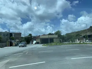 Bức ảnh chụp một con đường nhựa rộng rãi vào ban ngày, có vạch kẻ đường rõ ràng. Bầu trời trong xanh và có nhiều mây trắng. Một chiếc xe ô tô bán tải màu bạc đang di chuyển trên đường. Bên đường là các tòa nhà, bao gồm một garage sửa chữa ô tô với biển hiệu màu đỏ ghi "GARAGE Ô TÔ". Bên cạnh đó là một quán ăn có biển hiệu màu vàng ghi các món ăn, đồ uống: "BÁNH MÌ PATE", "BÁNH MỲ BÒ KHO", "XÔI", "BIA HƠI", "ĐỒ NHẬU". Phía sau các công trình là một ngọn đồi đá. Khung cảnh cho thấy đây là một khu vực giao thông ven đô thị hoặc thị trấn.。（日本語訳）。写真は、日中の、車線がはっきりと引かれた広々としたアスファルトの道路を写しています。空は青く澄み、白い雲が多く浮かんでいます。一台の銀色のピックアップトラックが道路を走行しています。道端には複数の建物があり、その中には「GARAGE Ô TÔ」と書かれた赤い看板を掲げた自動車修理工場が含まれています。その隣には、黄色い看板に「BÁNH MÌ PATE」、「BÁNH MỲ BÒ KHO」、「XÔI」、「BIA HƠI」、「ĐỒ NHẬU」といった料理や飲み物が書かれた食堂があります。それらの建物の後ろには岩山があります。この光景は、ここが都市の郊外または町の交通のある地域であることを示しています。