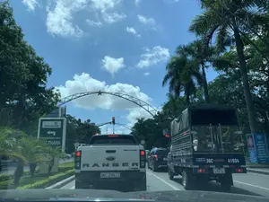 Bối cảnh là một con đường đô thị, có thể ở Thanh Hóa, được chụp từ bên trong một chiếc ô tô. Các phương tiện giao thông đang dừng chờ đèn đỏ dưới một cổng chào bằng kim loại có gắn ngôi sao vàng. Bầu trời trong xanh, có nắng và mây trắng, hai bên đường trồng nhiều cây xanh và cây cọ.。Hoạt động chính là các phương tiện đang tham gia giao thông và dừng lại theo tín hiệu đèn. Phía trước là một chiếc xe bán tải Ford Ranger màu trắng, bên phải là một chiếc xe tải có mui bạt.。Các vật thể và chữ viết được ghi nhận:。- Biển chỉ dẫn bên trái đường: 'Vietcombank', 'PHÒNG GIAO DỊCH TÁC THÀNH' và một mũi tên chỉ hướng.。- Trên xe tải bên phải: 'Ô TÔ THUẬN PHÁT', số điện thoại '0901.87.30.30', và 'Website: otothuanphat.vn'.。- Trên xe bán tải phía trước: 'Ford', 'RANGER', 'WILDTRAK'.。- Biển tuyên truyền màu xanh bên phải đường có nội dung: 'THAM GIA BẢO HIỂM XÃ HỘI TỰ NGUYỆN LÀ GÓP PHẦN GIẢM BỚT GÁNH NẶNG CHO GIA ĐÌNH VÀ XÃ HỘI KHI HẾT TUỔI LAO ĐỘNG' và 'THAM GIA BẢO HIỂM Y TẾ ĐỂ BẢO VỆ SỨC KHỎE CHO CÁC THÀNH VIÊN...'.。- Đèn tín hiệu giao thông đang hiển thị số đếm ngược là '16'.。（日本語訳）。背景は、おそらくタインホア省にある都市の道路で、自動車の中から撮影されたものです。交通車両は、黄色い星が取り付けられた金属製の歓迎ゲートの下で、赤信号で停止しています。空は青く澄み、晴れて白い雲があり、道路の両側には多くの緑の木々とヤシの木が植えられています。主な活動は、交通に参加している車両が信号に従って停止していることです。前方には白いフォード・レンジャーのピックアップトラックが、右側には幌付きのトラックがいます。確認された物体と文字：。- 道路左側の案内標識：「Vietcombank」、「タックタイン取引所」、および方向を示す矢印。- 右側のトラックに：「トゥアンファット自動車」、電話番号「0901.87.30.30」、および「ウェブサイト：otothuanphat.vn」。- 前方のピックアップトラックに：「Ford」、「RANGER」、「WILDTRAK」。- 道路右側の青いプロパガンダ看板の内容：「任意社会保険への加入は、労働年齢を終えた際の家族と社会への負担を軽減することに貢献します」および「医療保険に加入して、各メンバーの健康を守りましょう...」。- 交通信号機はカウントダウンの数字「16」を表示しています。