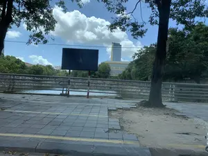 Bức ảnh ghi lại một khung cảnh đô thị bên cạnh một con kênh hoặc sông nhỏ vào một ngày nắng. Ở phía trước là vỉa hè lát gạch, một phần đã bị hư hỏng. Một lan can kim loại chạy dọc theo bờ kênh. Phía bên kia kênh là cây cối xanh tươi và các tòa nhà, trong đó có một tòa nhà cao tầng hiện đại. Có một biển báo hình chữ nhật màu sẫm trên vỉa hè, nhưng chữ viết trên đó đã mờ và không thể đọc được. Bầu trời trong xanh và có nhiều mây trắng.。（日本語訳）。写真は、晴れた日の、運河または小さな川の隣にある都市の風景を記録したものです。手前にはレンガ敷きの歩道があり、その一部は損傷しています。金属製の手すりが運河の岸に沿って走っています。運河の向こう側には、青々とした木々と建物があり、その中には近代的な高層ビルも含まれています。歩道には暗い色の長方形の看板がありますが、その上の文字は薄れていて読むことができません。空は青く澄んでおり、白い雲が多くあります。