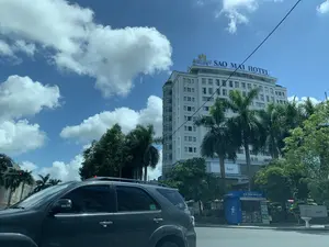 Bối cảnh là một góc phố đô thị vào một ngày nắng đẹp, trời xanh và có nhiều mây trắng. Nổi bật trong ảnh là tòa nhà cao tầng màu trắng của khách sạn Sao Mai, với biển hiệu lớn trên nóc ghi chữ "SAO MAI HOTEL". Phía trước, một chiếc ô tô SUV màu xám đang di chuyển hoặc dừng trên đường. Dọc theo con đường có nhiều cây xanh, bao gồm cả những cây cọ. Ở phía bên phải, có một ki-ốt màu xanh ghi "KI ỐT THÔNG TIN DU LỊCH" và một biển hiệu của "CoopBank". Trên tầng trệt của tòa nhà khách sạn cũng có một biển hiệu ghi chữ "Bank". Có thể thấy đèn tín hiệu giao thông đang báo màu đỏ.。（日本語訳）。背景は晴れた良い日の都市の街角で、空は青く、白い雲が多く浮かんでいます。写真の中で目立っているのはサオマイホテルの白い高層ビルで、屋上には「SAO MAI HOTEL」と書かれた大きな看板があります。手前では、灰色のSUV車が道路を走行しているか、停車しています。道沿いには多くの緑の木々があり、その中にはヤシの木も含まれています。右側には、「KI ỐT THÔNG TIN DU LỊCH」と書かれた青いキオスクと、「CoopBank」の看板があります。ホテルの建物の1階にも、「Bank」と書かれた看板があります。交通信号機が赤信号を示しているのが見えます。