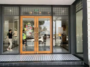 Bức ảnh chụp lối vào của một quán cà phê thuộc chuỗi 'The Coffee House'. Các thông tin bằng chữ có thể đọc được bao gồm: tên thương hiệu 'THE COFFEE HOUSE' được khắc dọc trên tay nắm cửa bằng gỗ; một tấm áp phích quảng cáo đồ uống với dòng chữ 'TƯƠI THẬT THẬT TƯƠI' và hình ảnh các loại trái cây; biển báo 'EXIT LỐI THOÁT' màu xanh lá cây ở phía trên cửa; và thông báo 'GIỜ MỞ CỬA: 7.30AM - 10.30PM'.。Về khung cảnh, đây là mặt tiền của một quán cà phê hiện đại với cửa kính lớn và sàn lát gạch hoa văn đen trắng. Bên trong quán, một người phụ nữ mặc áo đen đang đứng nói chuyện điện thoại. Phản chiếu trên cửa kính là khung cảnh đường phố bên ngoài với nhiều ô tô, một chiếc xe buýt màu vàng và các tòa nhà. Hoạt động chính là khách hàng ra vào quán và khung cảnh giao thông bên ngoài.。（日本語訳）。「ザ・コーヒーハウス」チェーンのカフェの入り口を撮影した写真です。読み取れる文字情報には、木製のドアハンドルに縦に刻まれたブランド名「THE COFFEE HOUSE」、飲み物を宣伝するポスターに書かれた「TƯƠI THẬT THẬT TƯƠI」（本当に新鮮）という文字と果物の画像、ドアの上にある緑色の「EXIT LỐI THOÁT」（出口）の標識、そして「GIỜ MỞ CỬA: 7.30AM - 10.30PM」（営業時間：午前7時30分～午後10時30分）という告知が含まれます。風景については、これは大きなガラスのドアと白黒の模様のタイルが敷かれた床を持つ、モダンなカフェの正面です。店内では、黒い服を着た女性が立って電話で話しています。ガラスのドアには、多くの自動車、一台の黄色いバス、そして建物が並ぶ外の通りの風景が映っています。主な活動は、客が店に出入りすることと、外の交通風景です。