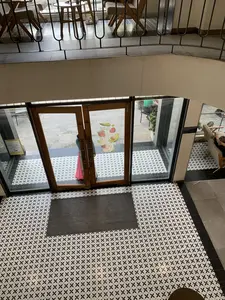 Bức ảnh được chụp từ trên cao nhìn xuống lối vào của một quán cà phê. Trên khung cửa kính bằng gỗ có dán dòng chữ "THE COFFEE HOUSE". Sàn nhà được lát bằng gạch hoa văn màu đen và trắng. Một người, dường như là một phụ nữ mặc áo dài màu hồng, đang đứng ở cửa. Qua cửa kính, có thể thấy không gian bên ngoài với bàn ghế và một vài khách hàng. Bên trong, ở tầng trên nơi chụp ảnh, có lan can bằng kim loại. Trên cửa kính còn có dán một poster quảng cáo đồ uống với hình ảnh các loại trái cây như dâu tây, bơ và mãng cầu. Có một tấm thảm chùi chân màu xám đặt trước cửa.。（日本語訳）。写真は、あるカフェの入り口を上から見下ろして撮影されたものです。木製のガラスドアのフレームには「THE COFFEE HOUSE」という文字が貼られています。床は黒と白の模様のタイルで敷き詰められています。一人の人物、ピンク色のアオザイを着た女性のようですが、ドアのところに立っています。ガラスドアを通して、テーブルと椅子、そして数人のお客さんがいる外の空間が見えます。内部、写真を撮っている上の階には、金属製の手すりがあります。ガラスドアには、イチゴ、アボカド、バンレイシといった果物の画像がある飲み物の広告ポスターも貼られています。ドアの前には灰色のドアマットが置かれています。