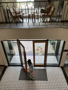 Bức ảnh được chụp từ trên cao nhìn xuống, bên trong một tòa nhà có vẻ là một quán cà phê. Ở tầng trên là khu vực ban công với lan can kim loại màu đen. Một người phụ nữ đang ngồi trên ghế gỗ, sử dụng điện thoại thông minh. Trên chiếc bàn tròn nhỏ bên cạnh cô là những ly đồ uống. Nhìn qua cửa sổ, có thể thấy khung cảnh đường phố bên ngoài. Ở tầng trệt là lối ra vào chính. Một người phụ nữ mặc áo dài truyền thống Việt Nam với họa tiết hoa sen và đội mũ đen đang bước qua cửa. Cửa ra vào làm bằng kính với khung gỗ, trên khung cửa có dòng chữ "THE COFFEE HOUSE" viết dọc. Trên cửa kính có dán một tấm áp phích với hình ảnh các loại trái cây như dâu tây, bơ, và mãng cầu. Sàn nhà được lát bằng gạch hoa văn màu đen và trắng.。（日本語訳）。写真は、カフェと思われる建物の中を、上から見下ろすように撮られたものです。上の階は、黒い金属製の手すりがあるバルコニーエリアです。一人の女性が木製の椅子に座り、スマートフォンを使っています。彼女の隣にある小さな丸いテーブルの上には、ドリンクのグラスがあります。窓からは、外の街の景色が見えます。1階はメインの出入り口です。蓮の花の模様が入ったベトナムの伝統的なアオザイを着て、黒い帽子をかぶった女性が、ドアを通り抜けています。出入り口のドアは木製のフレームが付いたガラス製で、ドアフレームには「THE COFFEE HOUSE」という文字が縦に書かれています。ガラスのドアには、イチゴ、アボカド、カスタードアップルのような果物の写真が載ったポスターが貼られています。床は、黒と白の模様付きタイルで敷き詰められています。