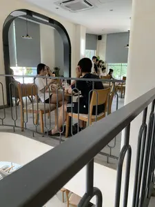 Đây là khung cảnh bên trong một quán cà phê có thiết kế hiện đại, có thể có nhiều tầng. Bức ảnh được chụp từ tầng trên nhìn xuống, qua một lan can kim loại màu xám. Ở khu vực trung tâm, một cặp đôi nam nữ đang ngồi tại một chiếc bàn gỗ. Người phụ nữ mặc áo phông trắng và quần short đen, đang nhìn vào điện thoại di động và mỉm cười. Người đàn ông mặc áo phông đen và quần short đen, quay lưng về phía máy ảnh, dường như đang trò chuyện. Trên bàn của họ có hai ly đồ uống bằng nhựa. Ở phía sau, có những khách hàng khác đang ngồi và đứng. Không gian quán có tường màu trắng, các ô cửa hình vòm, và cửa sổ lớn có rèm cuốn màu xám. Trên trần nhà có lắp đặt máy điều hòa không khí. Hoạt động chung là khách hàng đang thư giãn và giao lưu trong quán cà phê. Trên lưng áo của người đàn ông có dòng chữ nhưng không thể đọc rõ toàn bộ, có thể thấy chữ 'STREET'.。（日本語訳）。これは、モダンなデザインで、おそらく複数階建てのカフェの内部の光景です。写真は、灰色の金属製の手すり越しに、上の階から見下ろす形で撮影されています。中央エリアでは、一組の男女のカップルが木製のテーブルに座っています。女性は白いTシャツと黒いショートパンツを着ており、携帯電話を見て微笑んでいます。男性は黒いTシャツと黒いショートパンツを着てカメラに背を向けており、会話をしているようです。彼らのテーブルの上には、プラスチック製の飲み物のカップが2つあります。後方には、他の客が座ったり立ったりしています。店内の空間は、白い壁、アーチ型の開口部、そして灰色のロールスクリーンが付いた大きな窓があります。天井にはエアコンが設置されています。全体的な活動としては、客がカフェの中でリラックスし、交流しています。男性のTシャツの背中には文字がありますが、全てをはっきりと読み取ることはできず、「STREET」という文字が見えます。