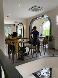 Trên lưng áo phông màu vàng của một người đàn ông có dòng chữ 'KỸ SƯ THANH HÓA' và số '77'. Bối cảnh là không gian bên trong một quán cà phê có thiết kế hiện đại, gồm nhiều tầng. Nhiều khách hàng đang ngồi tại các bàn gỗ, uống nước và trò chuyện. Một người đàn ông mặc áo polo màu xanh đang đứng và nói chuyện điện thoại. Quán có tường màu trắng, các cửa sổ lớn hình vòm, và được trang bị máy điều hòa âm trần. Bức ảnh được chụp từ tầng trên nhìn xuống khu vực tầng dưới, được ngăn cách bởi lan can kim loại. Có một chậu cây lưỡi hổ được đặt gần lan can để trang trí.。（日本語訳）。ある男性の黄色いTシャツの背中には「KỸ SƯ THANH HÓA」という文字と「77」という数字がある。背景は、モダンなデザインの多層階からなるカフェの店内空間である。多くの客が木製のテーブルに座り、飲み物を飲んだりおしゃべりをしたりしている。青いポロシャツを着た男性が立って電話で話している。写真は上の階から、金属製の手すりで仕切られた下の階のエリアを見下ろすように撮影されている。