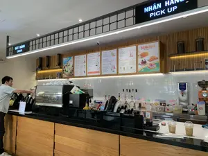 Bối cảnh là một quầy pha chế bên trong một quán cà phê hiện đại, có thể là một chi nhánh của The Coffee House dựa trên logo trên bảng menu. Một nhân viên nam đang đứng sau quầy, thao tác với máy pha cà phê chuyên nghiệp hiệu Simonelli. Quầy pha chế có mặt đá màu đen và thân ốp gỗ. Trên tường phía sau là gạch men trắng và các bảng menu đồ uống có đèn chiếu sáng. Hai ly cà phê sữa đá đã được pha xong, đặt trong ly nhựa và đang chờ được phục vụ.。Các thông tin bằng chữ viết được ghi lại:。- Biển hiệu chỉ dẫn: "ĐẶT HÀNG ORDER" và "NHẬN HÀNG PICK UP".。- Bảng menu đồ uống với các danh mục: "KHO MÀ BỐI", "CÀ PHÊ VIỆT NAM" (Đen/ Sữa/ Bạc Xỉu), "CÀ PHÊ SÁNG TẠO" (The Coffee House Sữa Đá, Đường Đen Sữa Đá, Đường Đen Marble Latte), "CÀ PHÊ MÁY" (Espresso/ Americano, Latte/ Cappuccino, Caramel Macchiato), "COLD BREW" (Truyền Thống, Sữa Tươi, Phúc Bồn Tử), "CLOUDTEA MOCHI" (Trà Xanh Tây Bắc | Oolong Berry), "TRÀ SỮA - MACCHIATO" (Trà Sữa Oolong B'Lao, Trà Sữa Oolong Nương Trân Châu, Trà Sữa Oolong Tử Quý Sương Sáo, Hồng Trà Sữa Trân Châu, Trà Đen Macchiato), "SÔ CÔ LA", "FROSTY" (Phin-Gato, Caramel Arabica, Cà Phê Đường Đen, Bánh Kem Dâu, Choco Chip), "TRÀ XANH TÂY BẮC" (Trà Xanh Latte, Trà Xanh Espresso Marble, Trà Xanh Đường Đen, Frosty Trà Xanh), "TRÀ TRÁI CÂY" (Trà Đào Cam Sả, Oolong Tử Quý Sen, Oolong Tử Quý Vải, Oolong Tử Quý Kim Quất Trân Châu), "HI-TEA HEALTHY" (Đào | Vải | Yuzu Trân Châu, Đào Kombucha | Yuzu Kombucha), "TRÁI CÂY XAY 0°C" (Bơ Sữa Phô Mai Tươi, Măng Cầu Phô Mai Tươi, Dâu Phô Mai Tươi, Mận Phô Mai Tươi, Nho Phô Mai Tươi).。- Bảng quảng cáo bánh: "BÁNH NHÀ - Mặn ngọt đủ đầy" với hình ảnh bánh mì chà bông, bánh sừng bò, bánh gấu sô cô la và mochi.。（日本語訳）。背景はモダンなカフェの中にあるバーカウンターで、メニューボードのロゴから「The Coffee House」の支店である可能性があります。一人の男性スタッフがカウンターの後ろに立ち、Simonelli製のプロ仕様のコーヒーマシンを操作しています。バーカウンターは天板が黒い石で、本体は木で覆われています。後ろの壁には白いタイルが貼られ、照明付きのドリンクメニューボードがあります。2杯のアイスミルクコーヒーが淹れ終わり、プラスチックカップに入れられて提供を待っています。記載されている文字情報：。- 案内看板：「ĐẶT HÀNG ORDER」（ご注文）と「NHẬN HÀNG PICK UP」（お受け取り）。- ドリンクメニューボードと各カテゴリー：「KHO MÀ BỐI」、「CÀ PHÊ VIỆT NAM」（ベトナムコーヒー）（ブラック／ミルク／バクシウ）、「CÀ PHÊ SÁNG TẠO」（クリエイティブコーヒー）（The Coffee House アイスミルクコーヒー、黒糖アイスミルクコーヒー、黒糖マーブルラテ）、「CÀ PHÊ MÁY」（マシンコーヒー）（エスプレッソ／アメリカーノ、ラテ／カプチーノ、キャラメルマキアート）、「COLD BREW」（コールドブリュー）（トラディショナル、フレッシュミルク、ラズベリー）、「CLOUDTEA MOCHI」（クラウドティー・モチ）（タイバック緑茶 | ウーロンベリー）、「TRÀ SỮA - MACCHIATO」（ミルクティー - マキアート）（バオロク・ウーロンミルクティー、ヌオン・ウーロンミルクティー・タピオカ入り、四季春ウーロンミルクティー・仙草ゼリー入り、紅茶ミルクティー・タピオカ入り、ブラックティー・マキアート）、「SÔ CÔ LA」（チョコレート）、「FROSTY」（フロスティ）（フィンガート、キャラメルアラビカ、黒糖コーヒー、ストロベリーケーキ、チョコチップ）、「TRÀ XANH TÂY BẮC」（タイバック緑茶）（緑茶ラテ、緑茶エスプレッソマーブル、緑茶黒糖、緑茶フロスティ）、「TRÀ TRÁI CÂY」（フルーツティー）（ピーチ・オレンジ・レモングラスティー、四季春ウーロン・蓮茶、四季春ウーロン・ライチ茶、四季春ウーロン・キンカン・タピオカ茶）、「HI-TEA HEALTHY」（ヘルシー・ハイティー）（ピーチ | ライチ | ユズ・タピオカ、ピーチコンブチャ | ユズコンブチャ）、「TRÁI CÂY XAY 0°C」（0°Cフルーツスムージー）（アボカド・ミルク・フレッシュチーズ、サワーソップ・フレッシュチーズ、ストロベリー・フレッシュチーズ、プラム・フレッシュチーズ、グレープ・フレッシュチーズ）。- ペストリーの広告ボード：「BÁNH NHÀ - Mặn ngọt đủ đầy」（自家製ペストリー - 甘いものからしょっぱいものまで充実）と書かれ、肉でんぶパン、クロワッサン、熊の形のチョコレートケーキ、餅の写真が掲載されています。