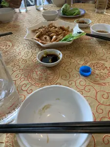 Bối cảnh là một bữa ăn đang diễn ra trên một chiếc bàn được trải khăn màu vàng với hoa văn màu đỏ. Món ăn chính là một đĩa tôm rang được bày trên đĩa sứ trắng hình ngôi sao, có trang trí rau thơm. Xung quanh là các vật dụng ăn uống như bát sứ trắng, đũa đen, và cốc nước thủy tinh. Có những bát nhỏ đựng nước chấm và một đĩa rau sống (xà lách). Ở phía trước, một chiếc bát đã vơi và một đôi đũa được đặt lên trên. Có một cái nắp chai nhựa màu xanh dương nằm trên bàn. Hình ảnh cho thấy mọi người đang dùng bữa.。（日本語訳）。背景は、赤い模様の入った黄色いテーブルクロスが敷かれたテーブルの上で、食事が行われている場面です。メインディッシュは、香草が飾られた、星形の白い磁器の皿に盛り付けられたエビの炒め物です。周りには、白い磁器の碗、黒い箸、ガラスのコップといった食器類があります。タレを入れた小鉢と、生野菜（レタス）の皿があります。手前には、中身が減った碗とその上に置かれた一膳の箸があります。テーブルの上には、青いプラスチックのボトルキャップが一つあります。この画像は、人々が食事をしていることを示しています。