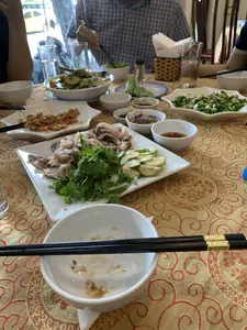 Đây là cảnh một bữa ăn tại một nhà hàng. Trên bàn ăn được trải một tấm khăn trải bàn màu vàng có hoa văn, bày biện nhiều món ăn Việt Nam. Có nhiều người đang cùng nhau dùng bữa. Các món ăn trên bàn bao gồm một đĩa lớn thịt luộc thái mỏng ăn kèm với rau thơm và ổi xanh hoặc vả thái lát. Ngoài ra còn có một đĩa tôm rang, một đĩa nộm (gỏi), và một bát canh. Nhiều bát nước chấm nhỏ như nước mắm ớt và có thể là mắm tôm được đặt trên bàn. Một người đàn ông mặc áo sơ mi kẻ caro đang dùng đũa để gắp thức ăn. Trên bàn còn có bát con, đũa, cốc thủy tinh và giỏ đựng giấy ăn.。（日本語訳）。これはレストランでの食事の風景です。食卓には、模様の入った黄色いテーブルクロスが敷かれ、多くのベトナム料理が並べられています。多くの人々が一緒に食事をしています。テーブルの上の料理には、香草やスライスした青いグアバまたはイチジクを添えた、薄切り茹で豚の大皿が含まれています。その他に、エビの炒め物一皿、ノム（和え物）一皿、そしてスープ一杯があります。唐辛子入りヌクマムや、おそらくマムトムのような、多くの小さなつけだれの小鉢がテーブルに置かれています。チェック柄のシャツを着た男性が、箸を使って食べ物を取っています。テーブルの上には、小碗、箸、ガラスのコップ、そして紙ナプキンを入れるかごもあります。