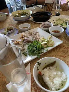 Bối cảnh là một bàn ăn tại một nhà hàng hoặc một bữa tiệc gia đình, có vẻ như bữa ăn đã gần kết thúc. Trên bàn được trải một chiếc khăn màu vàng có hoa văn. Có rất nhiều bát đĩa sứ trắng đã qua sử dụng với thức ăn còn sót lại. Các món ăn trên bàn bao gồm một đĩa lớn ở trung tâm có thịt luộc, tôm, rau thơm và một loại quả xanh thái lát (trông giống sung muối hoặc ổi xanh). Ngoài ra còn có một nồi đất màu đen đựng món kho, các bát nước chấm (một số có ớt, một số giống mắm tôm), và một bát cơm trắng với cá kho ở phía trước. Mọi người dường như đã dùng bữa xong. Có thể thấy các cốc thủy tinh, đũa, và một giỏ đựng giấy ăn. Trên giấy ăn có in chữ màu đỏ nhưng chỉ đọc được một phần, có vẻ là tên của một nhà hàng.。（日本語訳）。背景はレストランか家族のパーティーの食卓で、食事はもうすぐ終わりのようです。テーブルの上には模様のある黄色いテーブルクロスが敷かれています。使用済みで食べ残しのある白い陶器の皿や碗がたくさんあります。テーブルの上の料理には、中央に茹で肉、エビ、香草、そしてスライスされたある種の青い果物（塩漬けのイチジクか青いグアバのように見えます）が乗った大皿が含まれます。その他にも、煮込み料理が入った黒い土鍋、タレの入った小鉢（いくつかは唐辛子入り、いくつかはマムトムのようです）、そして手前には魚の煮付けが乗った白いご飯の碗があります。皆さんは食事を終えたようです。ガラスのコップ、箸、そして紙ナプキンを入れるかごが見えます。紙ナプキンには赤い文字が印刷されていますが、一部しか読めず、レストランの名前のようです。